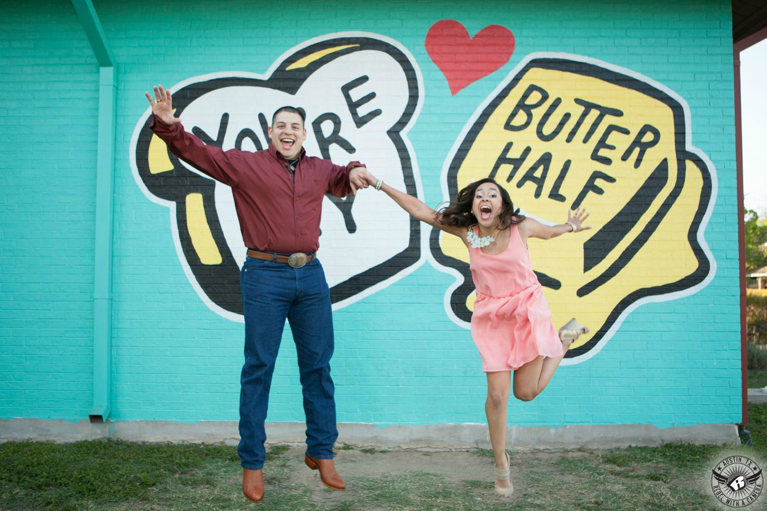 Latino girl with dark hair wearing a pink dress, strappy tan heels and a diamond necklace jumps for joy while holding the hand of a Latino guy with dark hair wearing a maroon button up shirt, a large belt buckle, blue jeans and cowboy boots also jumping in front of the You're My Butter Half mural in this fun engagement photo in East Austin